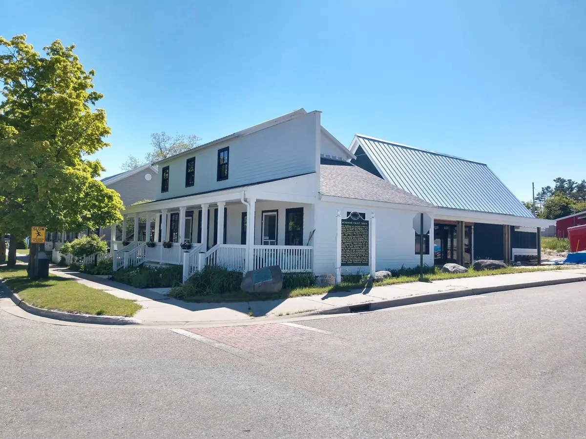 White two-story building with a covered porch, metal-roofed addition, roadside plaque, and warning sign.