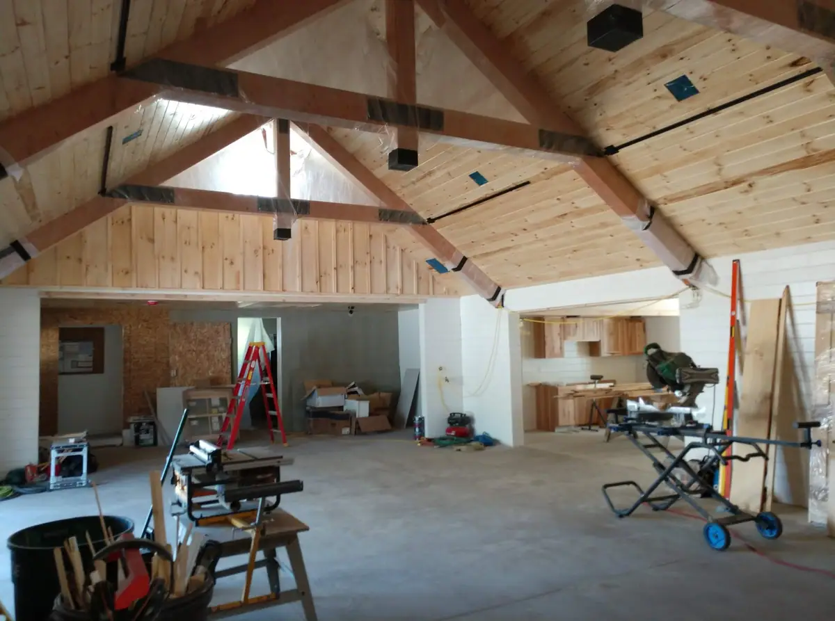 Interior of a large unfinished workshop with a vaulted wooden ceiling, tools, ladders, cabinets, and construction materials.