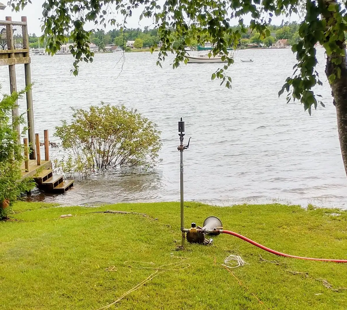 A flooded waterfront lawn with a wooden dock, metal fixture, hose, boats, and houses across the water.