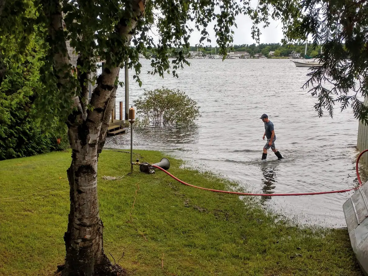 A person wades through high water beside a grassy waterfront, with a red hose, trees, and a dock visible.