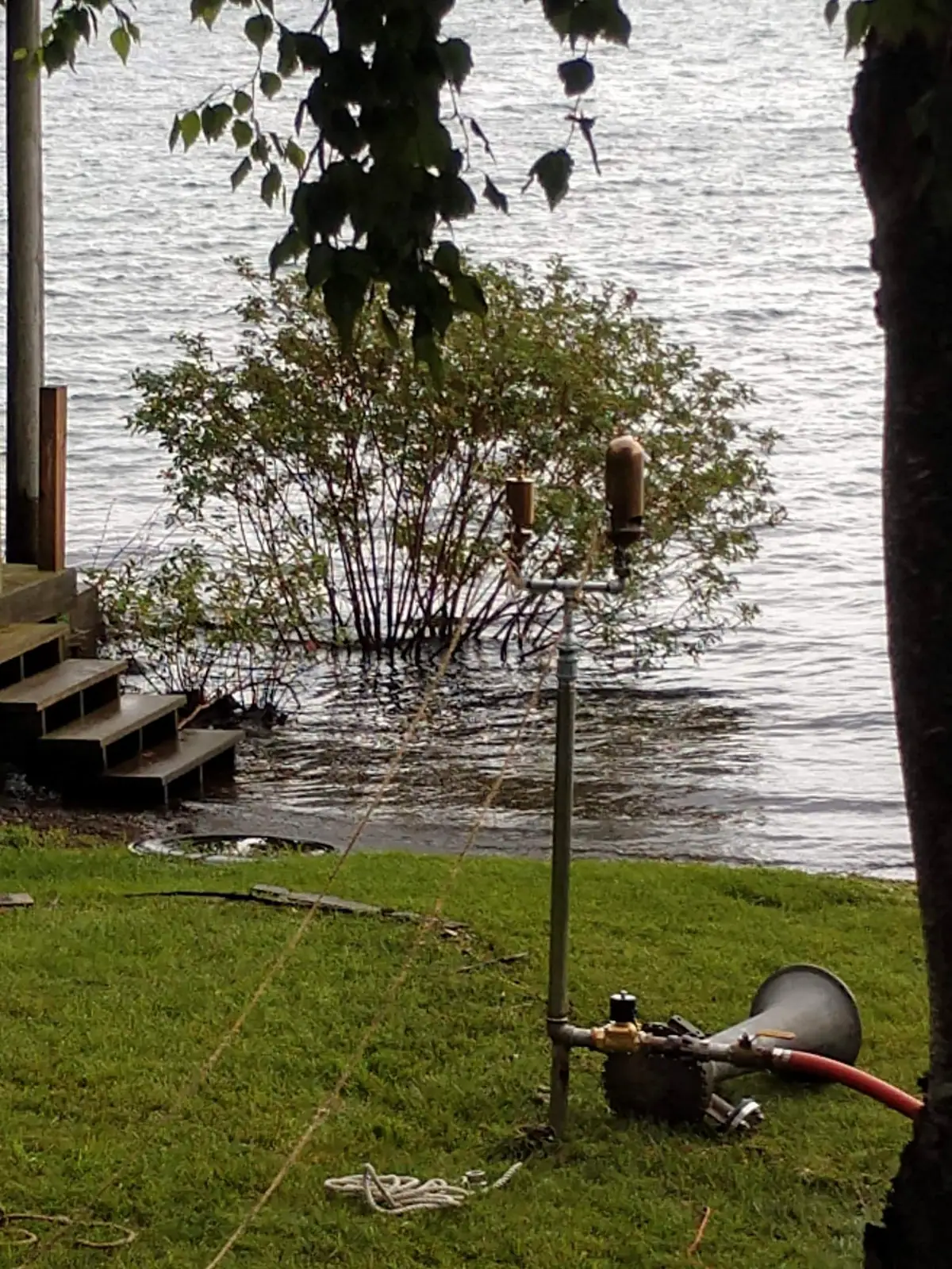 Floodwater surrounds a shrub beside wooden steps, with a hydrant assembly, hose, horn-shaped device, and rope in the foreground.