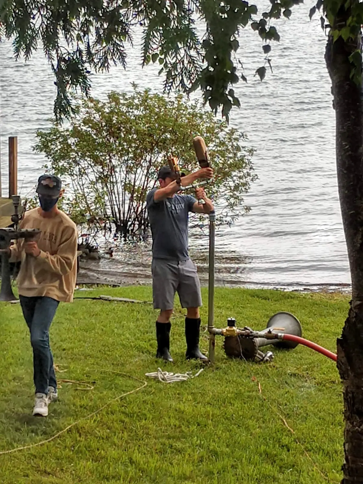 Two men work with a vertical pipe, hoses, and equipment on a grassy shoreline.