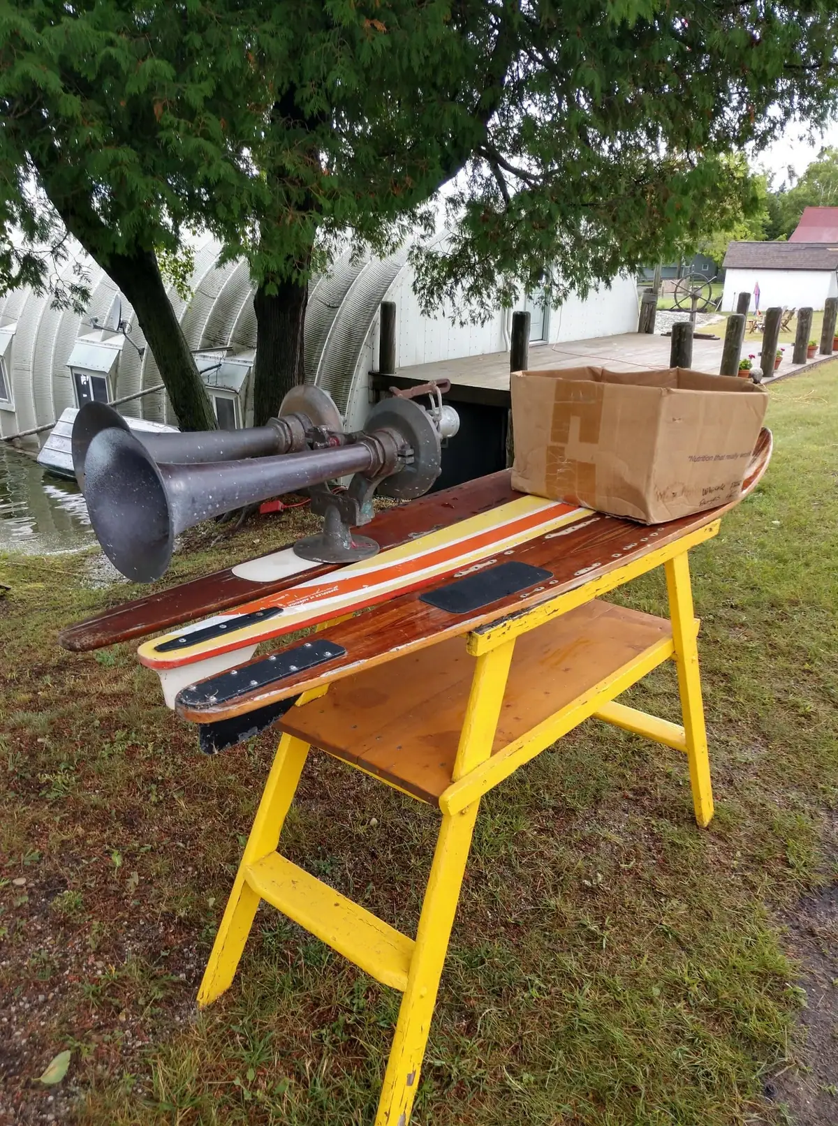 A yellow wooden table displays long wooden boards, a large metal horn, and a cardboard box outdoors.