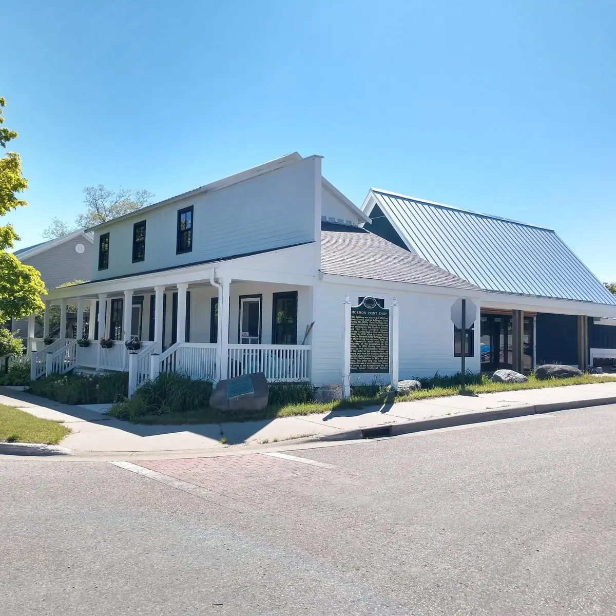 A white two-story building with a covered porch, metal-roofed addition, and an interpretive sign beside a paved street.