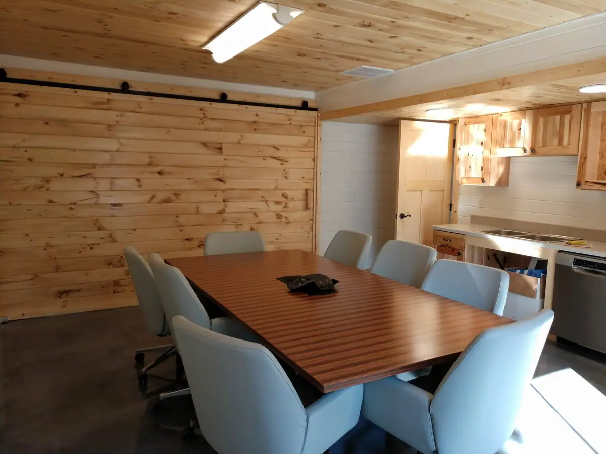 Wood-paneled meeting room with a large table, blue chairs, and a compact kitchenette.
