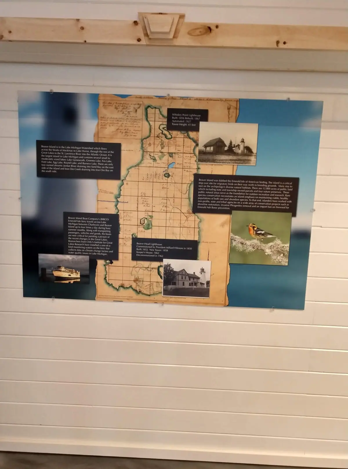 Photograph of a wall-mounted exhibit panel with a map, text, lighthouse images, a ferry, and a bird.