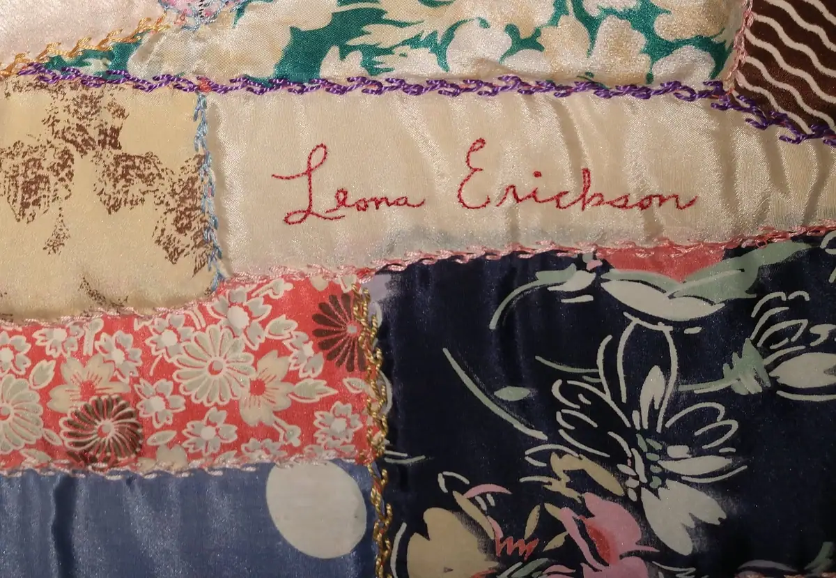 Close-up of a colorful patchwork textile with embroidered edging and the handwritten name “Lona Erickson.”