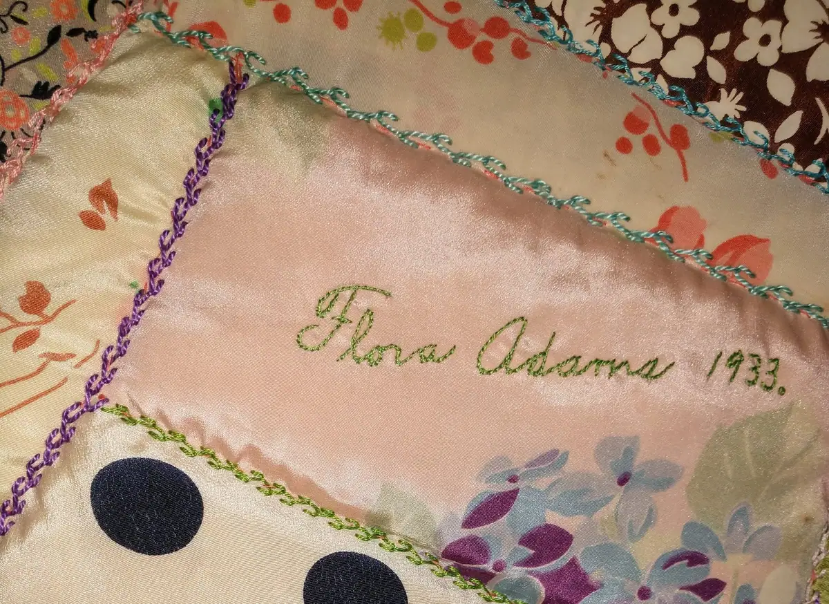 Close-up of a floral textile with multicolored stitched edging and embroidered lettering reading “Ida Adams 1933.”