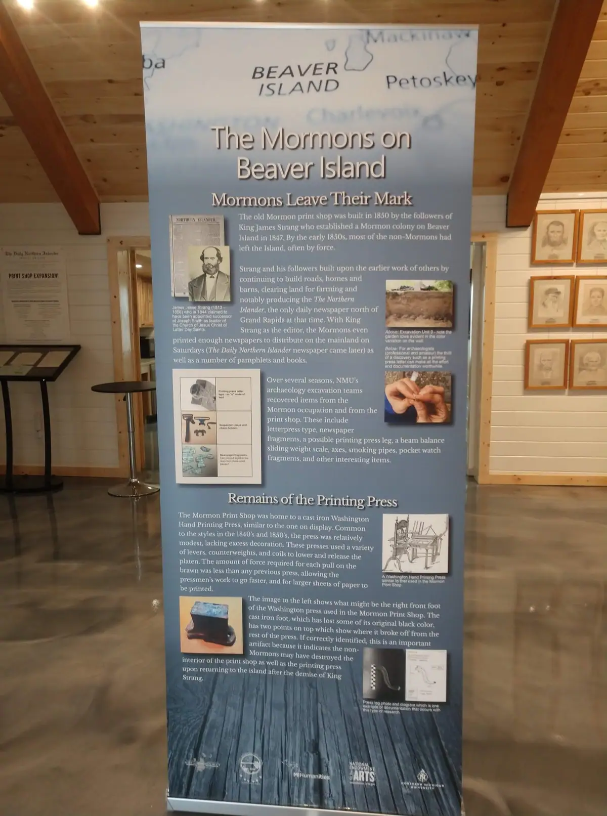 A tall exhibit banner about a historic printing press stands in a wood-paneled gallery beside framed portraits.