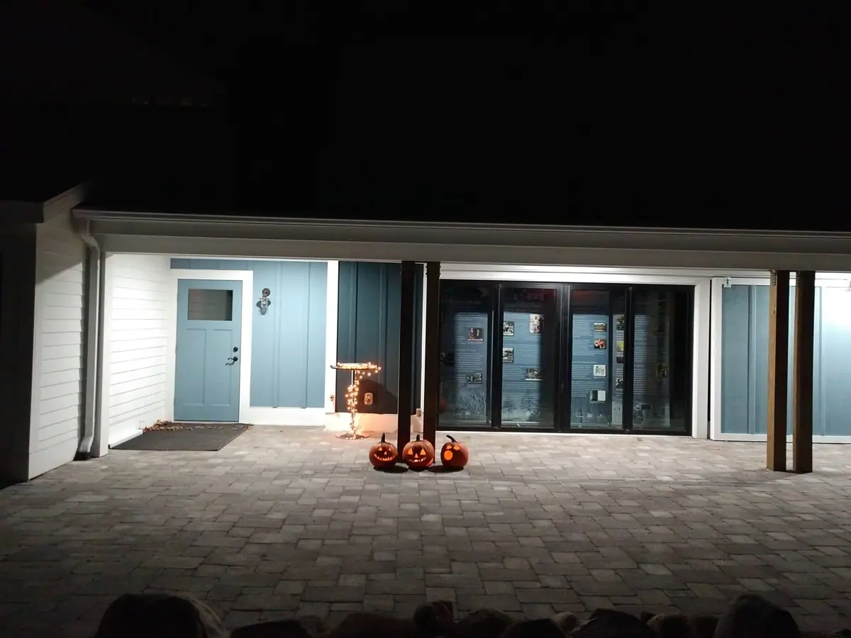 Night photograph of a covered building entrance with three glowing carved pumpkins on a paved walkway.