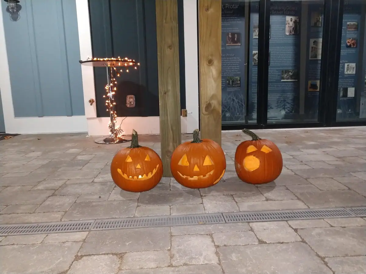 Three illuminated carved pumpkins sit on a paved patio outside a building with exhibit panels.