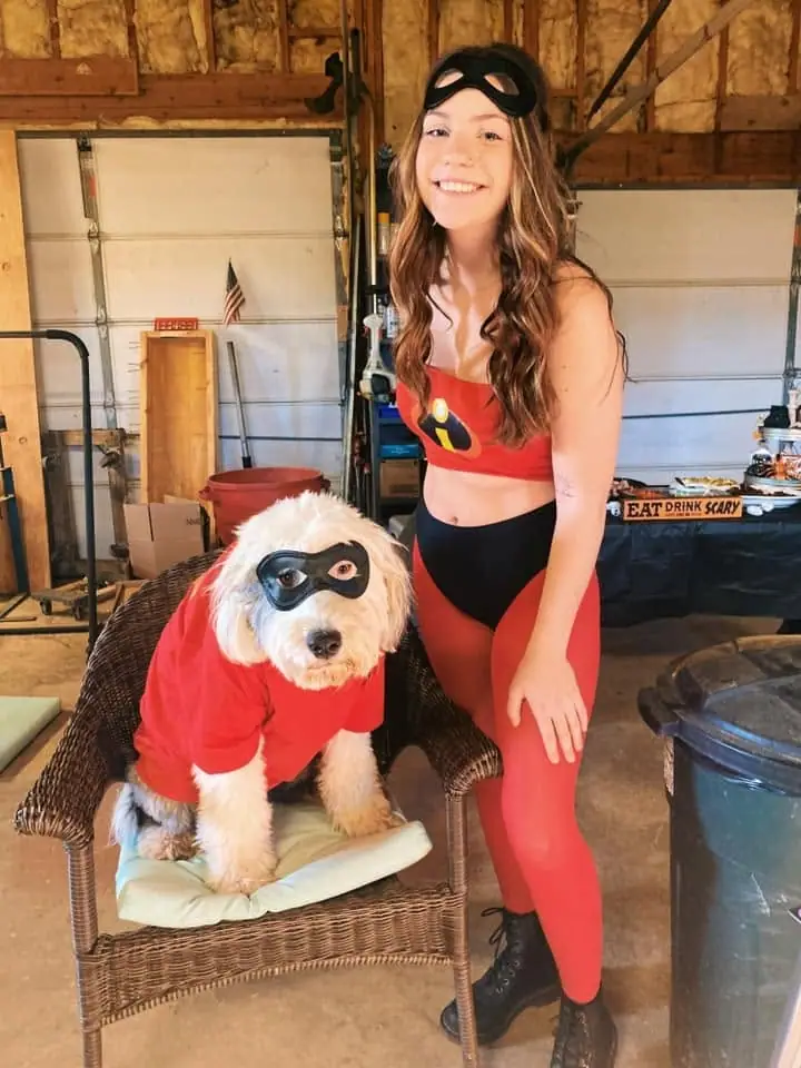 A woman and a masked dog pose in matching red superhero costumes inside a garage.