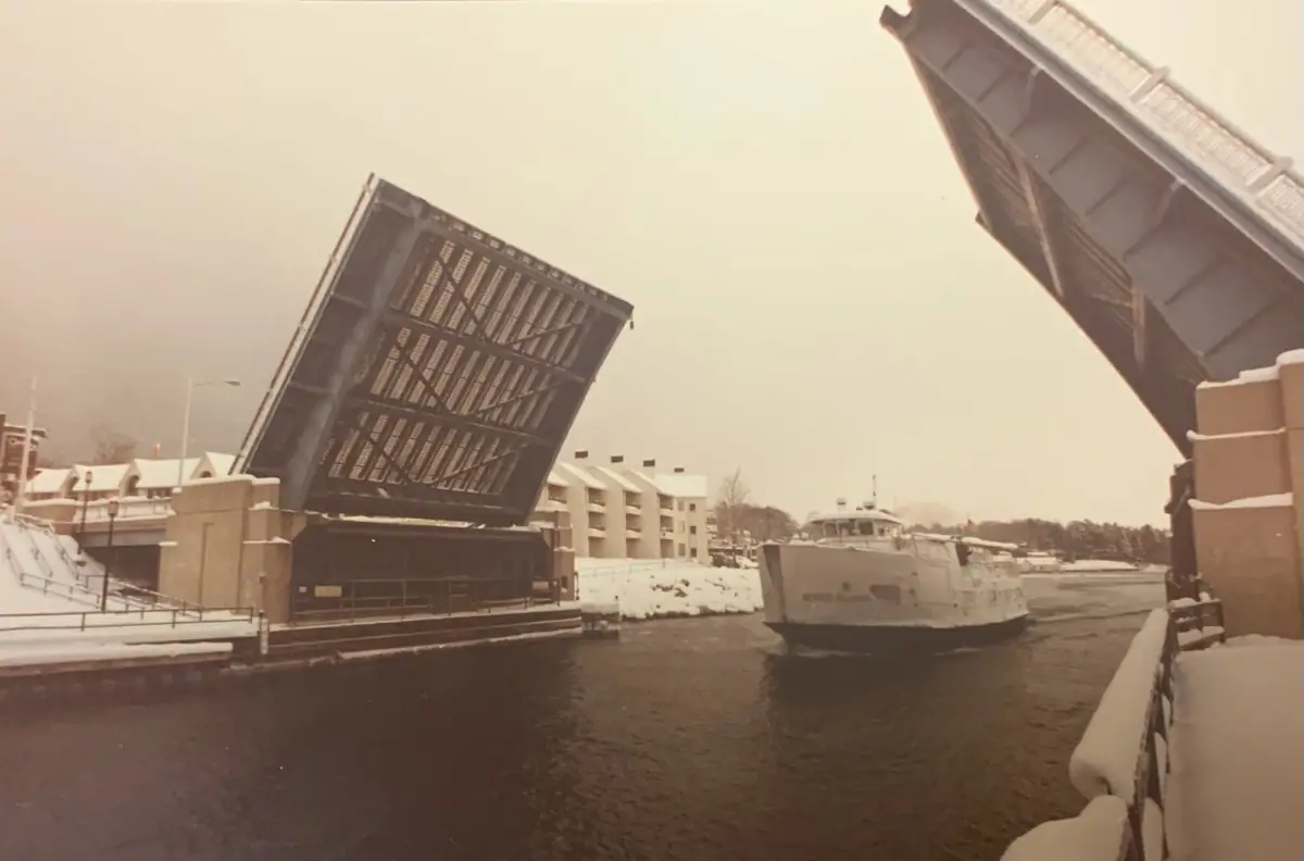 A white ferry passes beneath a raised drawbridge on a snowy winter waterway.