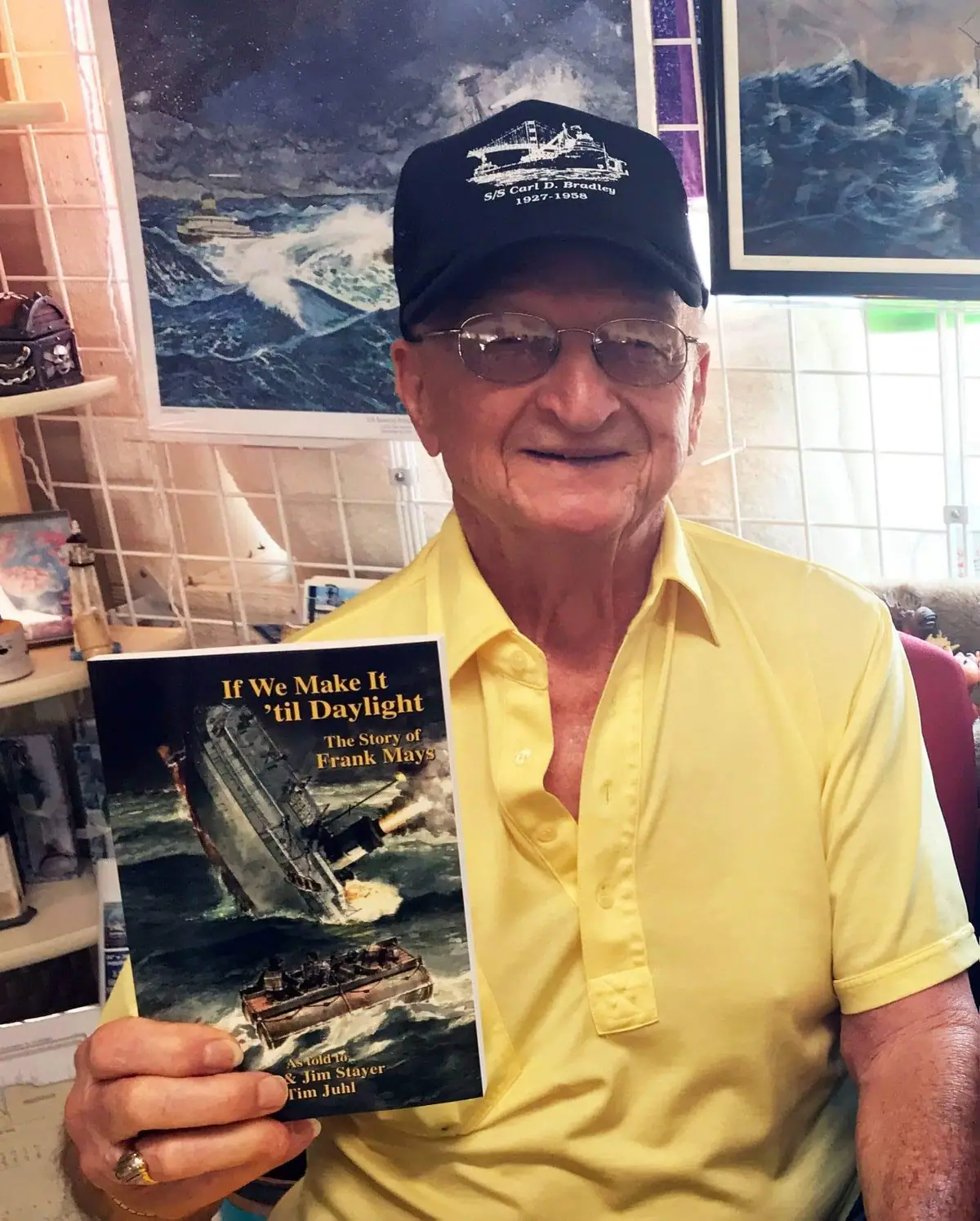 A man in a yellow shirt holds a book about a shipwreck while wearing a maritime baseball cap.