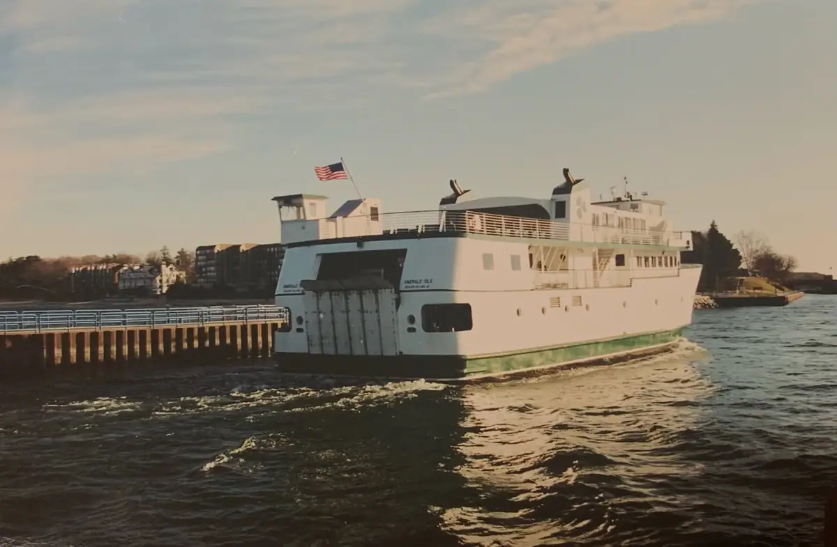 A white ferry with a green stripe passes a pier, leaving a wake beneath a partly cloudy sky.
