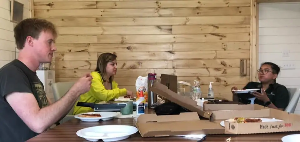 Three people share pizza at a table in a wood-paneled room.