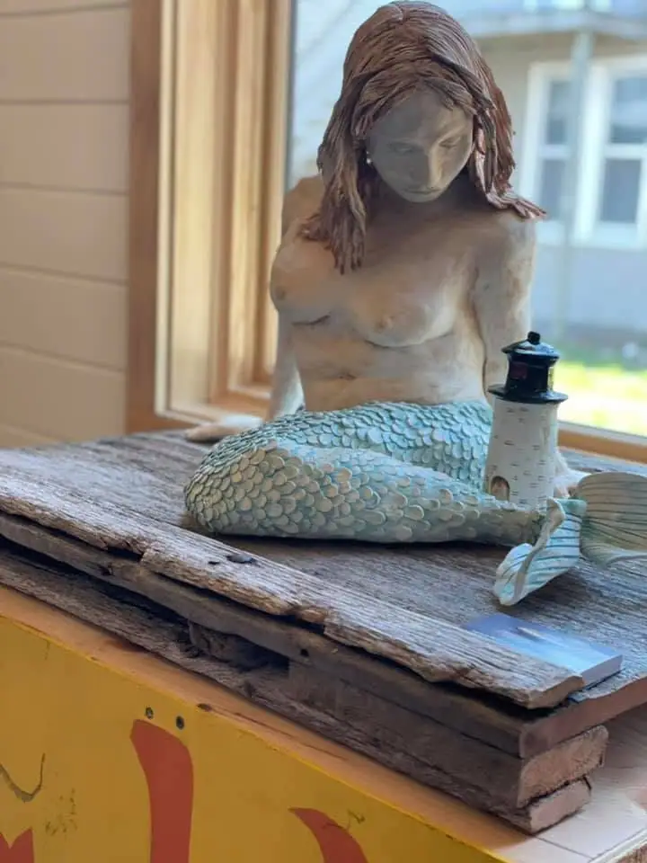 Photograph of a mermaid sculpture with a miniature lighthouse on a rough wooden platform.