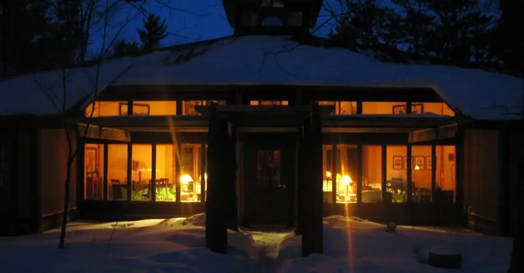 A snow-covered, low building glows with warm light through its large windows at dusk.