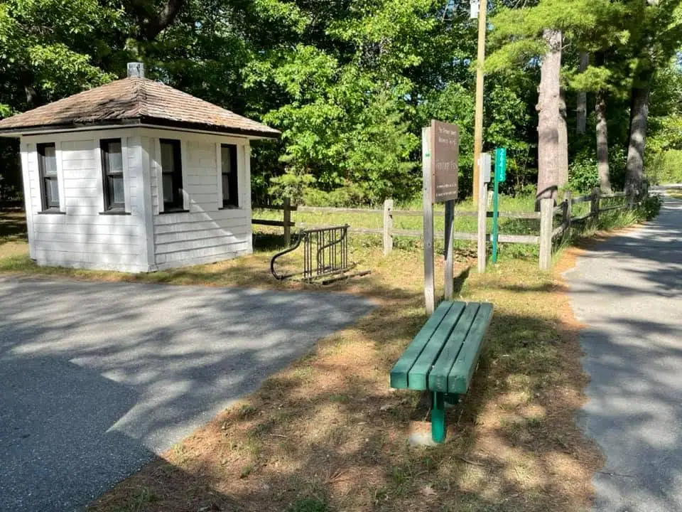 Small white wooden building beside a fenced path, bench, and signposts among trees.