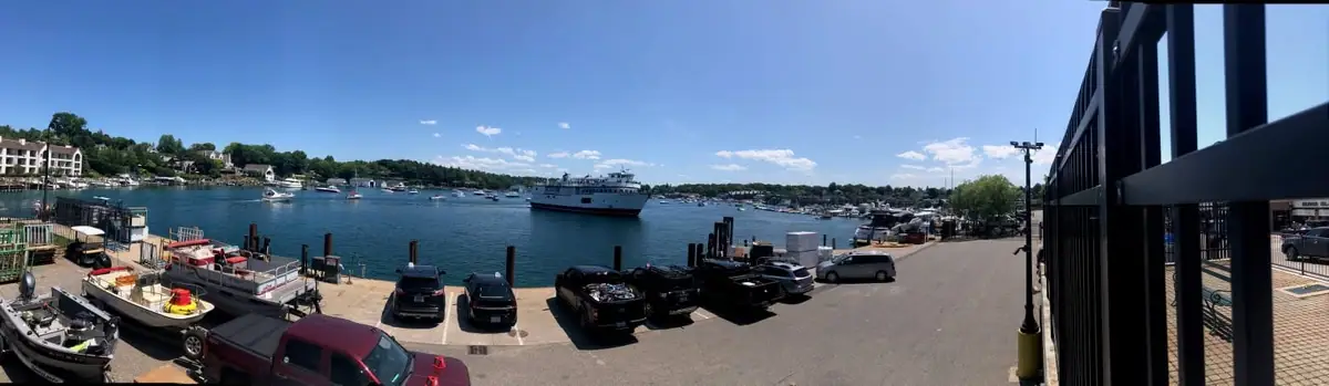 Panoramic waterfront view with a passenger ferry, small boats, docks, vehicles, and a large building.