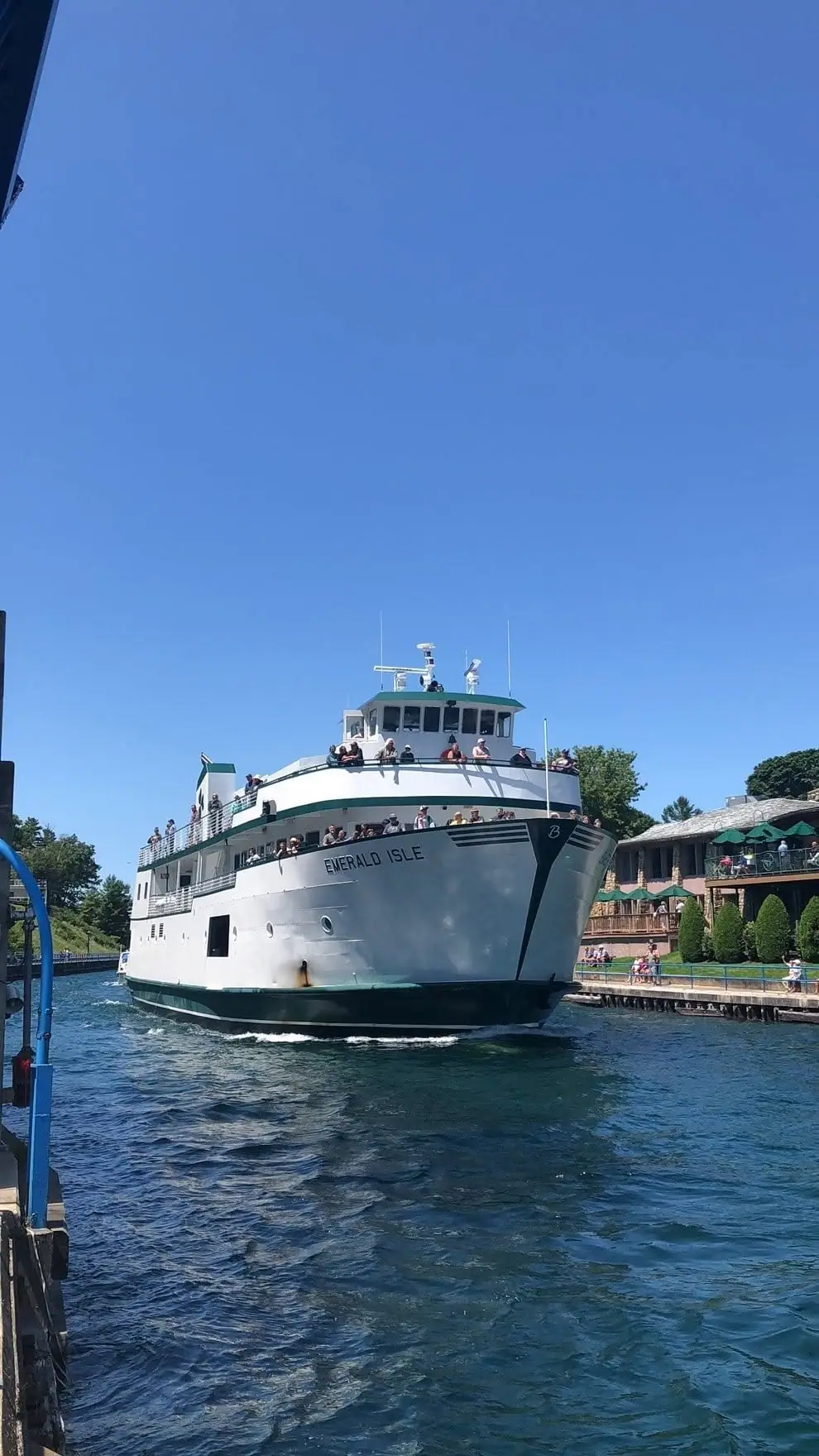 A large white passenger ferry approaches a dock, with people on its decks and along the waterfront.