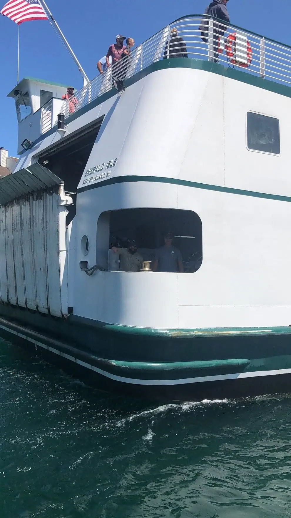 White-and-green ferry with passengers and an American flag, viewed close-up on the water beside a dock.