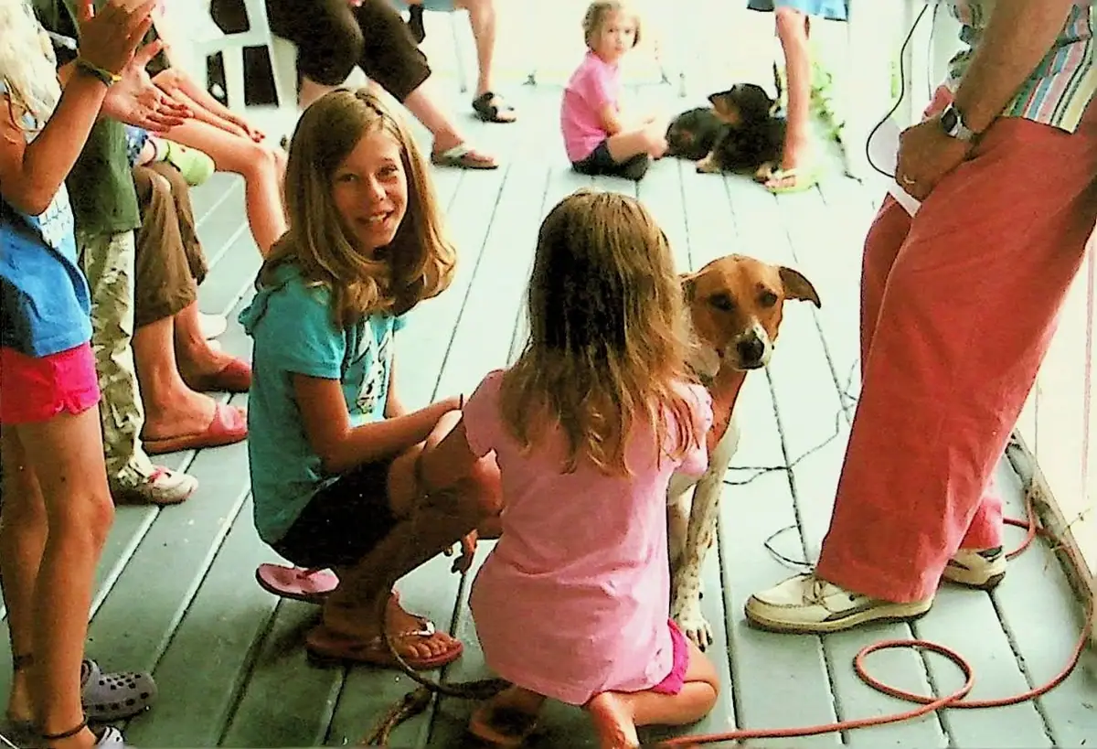 Children and adults gather around two dogs on a light-colored wooden deck.