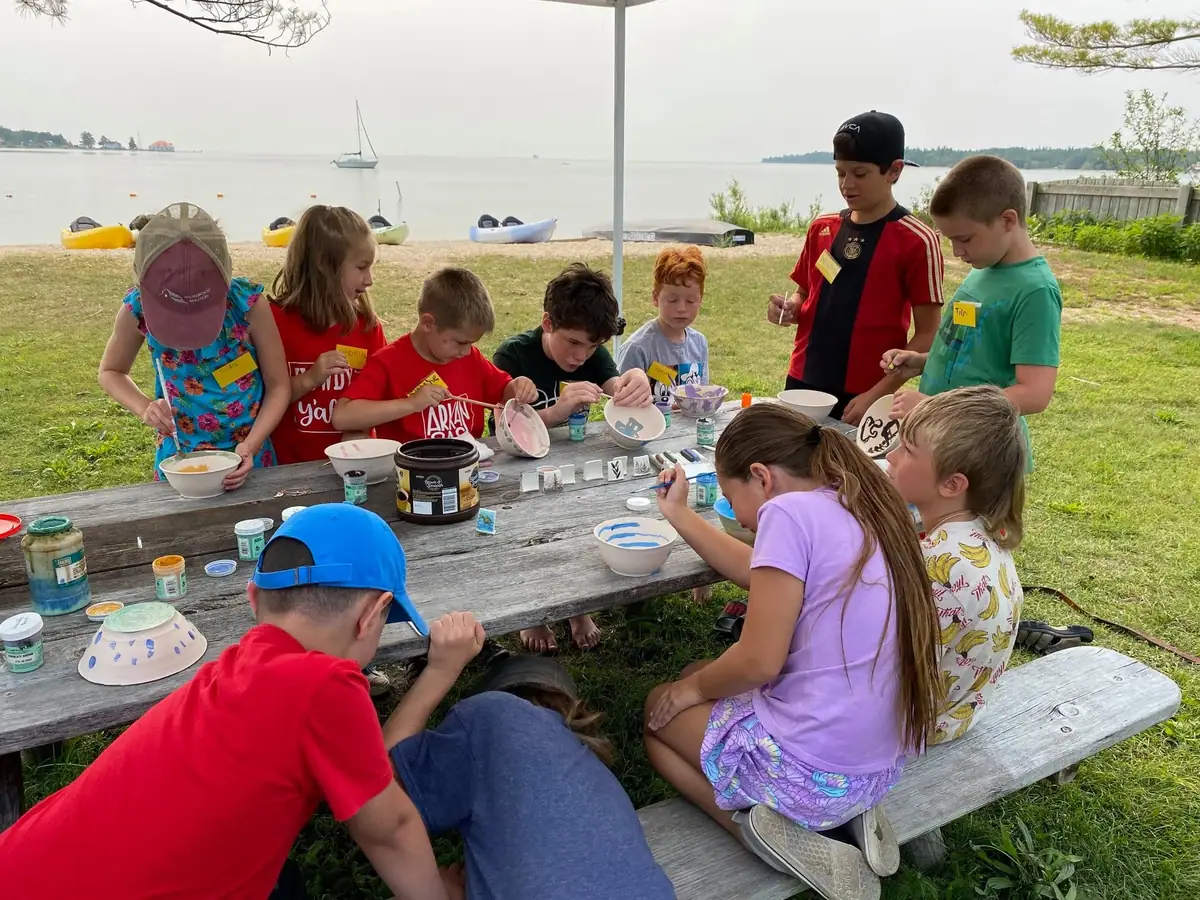 Children paint ceramic bowls and other pottery at a picnic table beside a shoreline, with boats visible behind them.