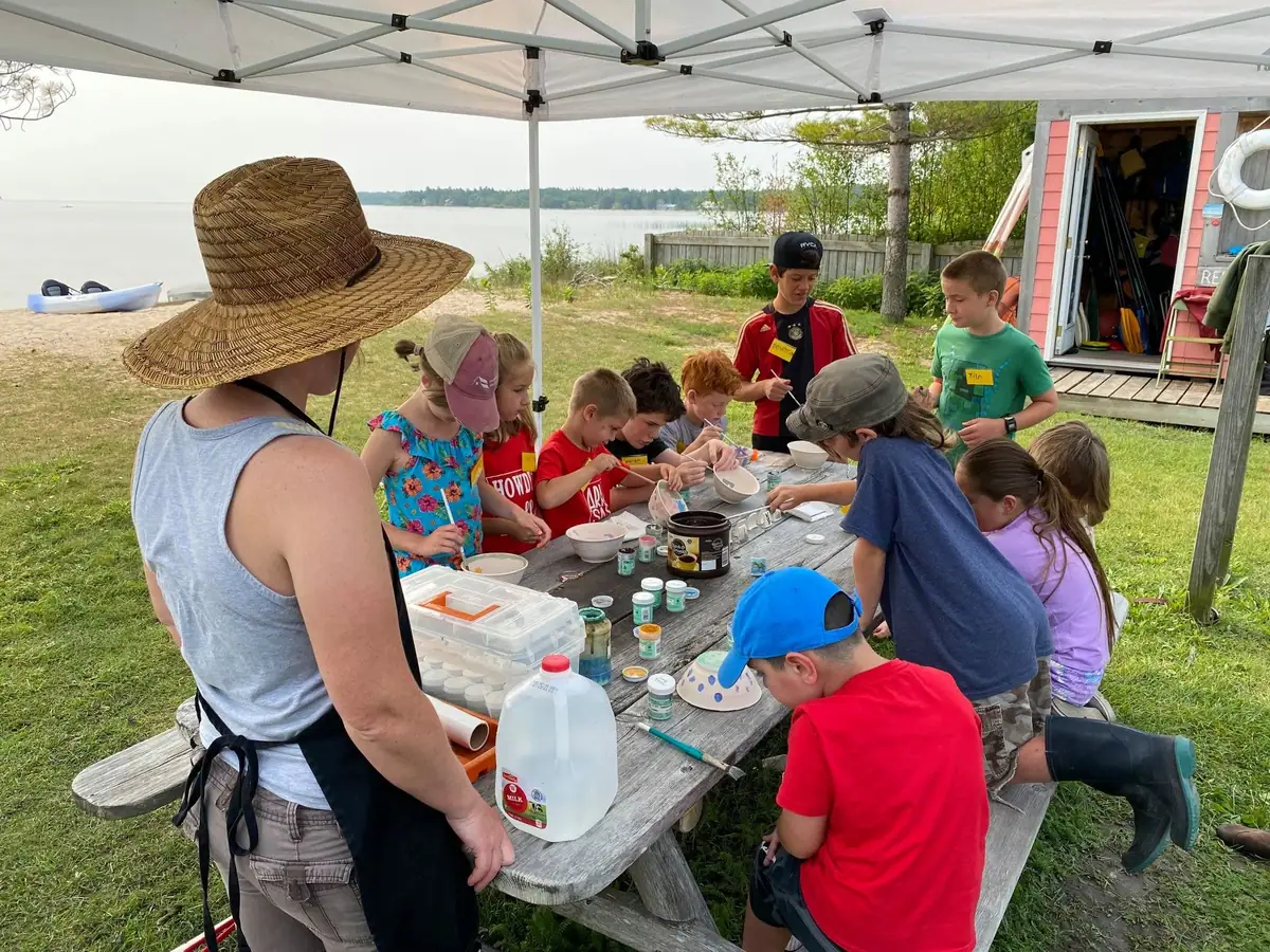 Children and adults paint small bowls at a picnic table beneath a canopy beside the shoreline.