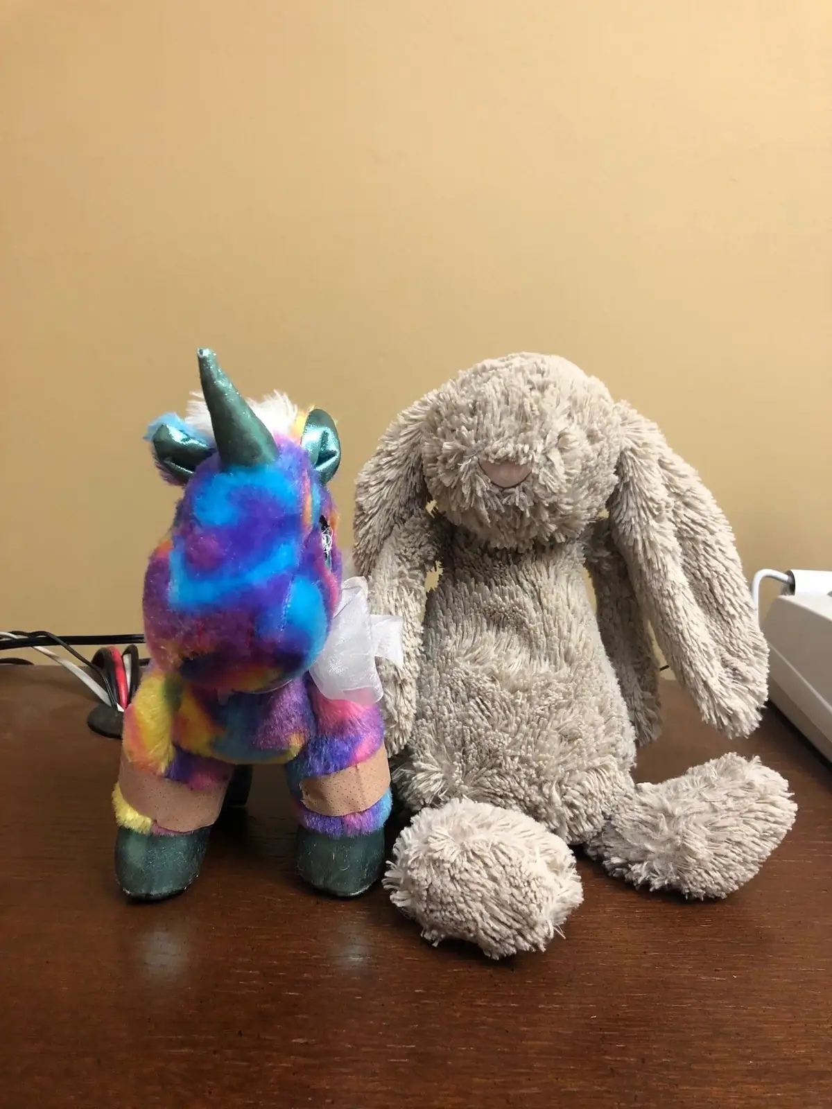 A colorful plush unicorn and a gray plush bunny sit side by side on a wooden tabletop.