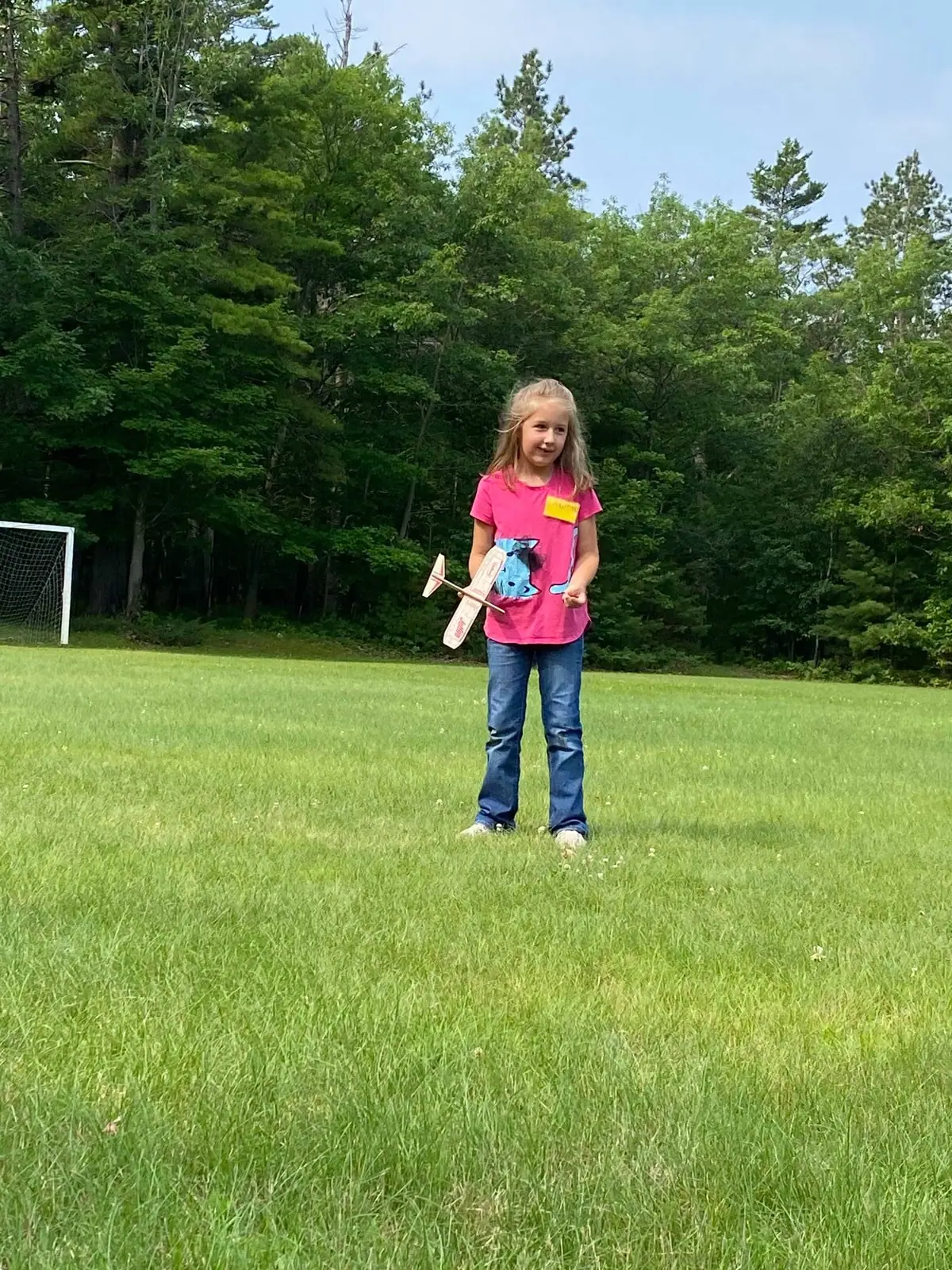 A child holds a small model airplane in a grassy field beside a soccer goal and forest.