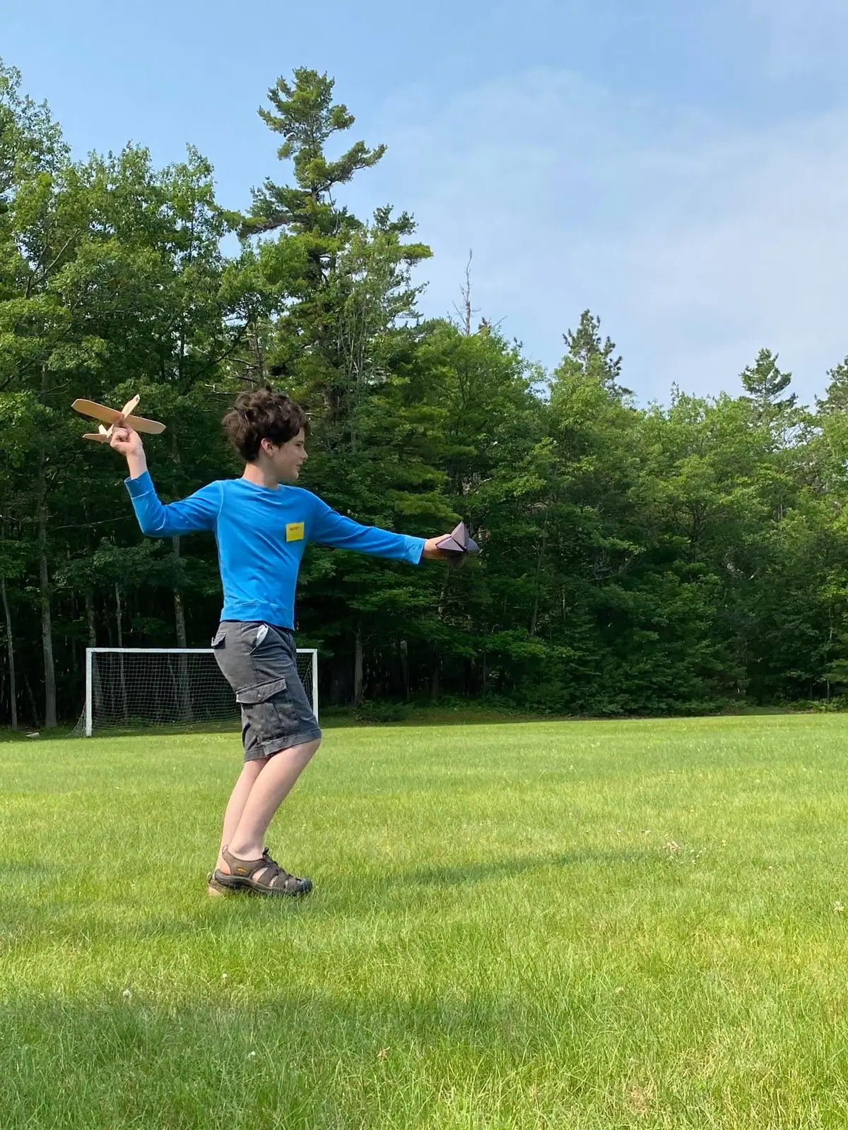 Child holding a wooden airplane and a paper airplane in a grassy field with a soccer goal and trees behind.