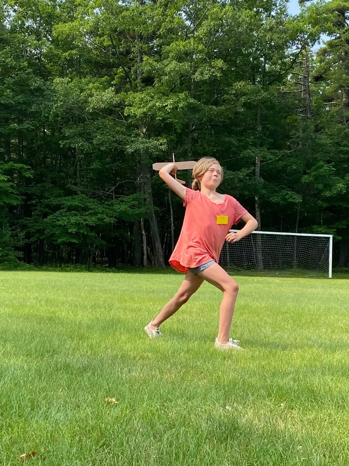 Child posing with a wooden airplane model on a grassy field beside a soccer goal.