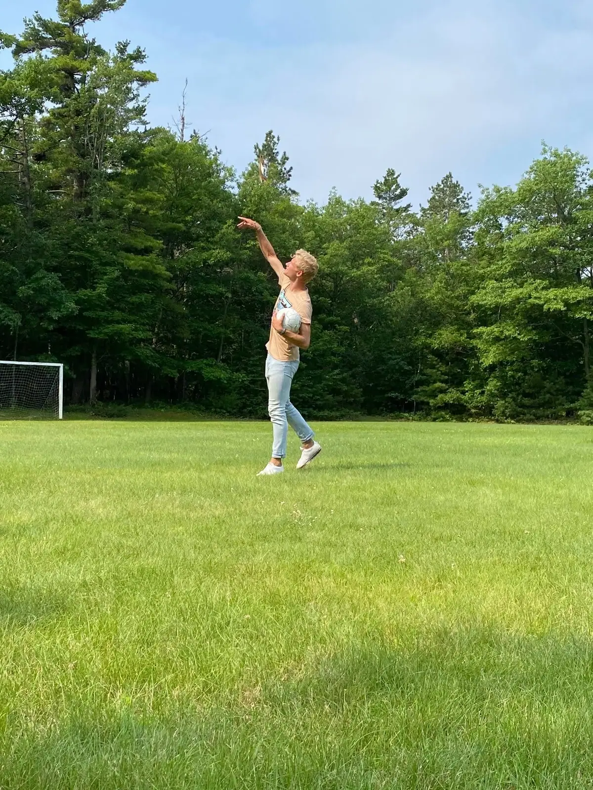 A person holding a soccer ball points upward on a grassy field beside a goal and forest.