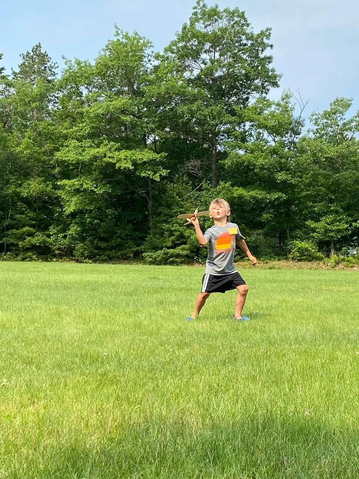 Child playing with a wooden model airplane in a grassy field bordered by trees.