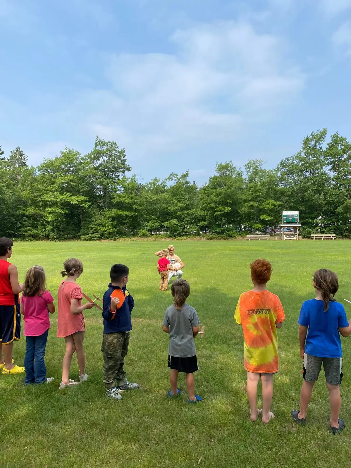 Children watch two people with a ball on a grassy field, with a scoreboard and trees behind them.