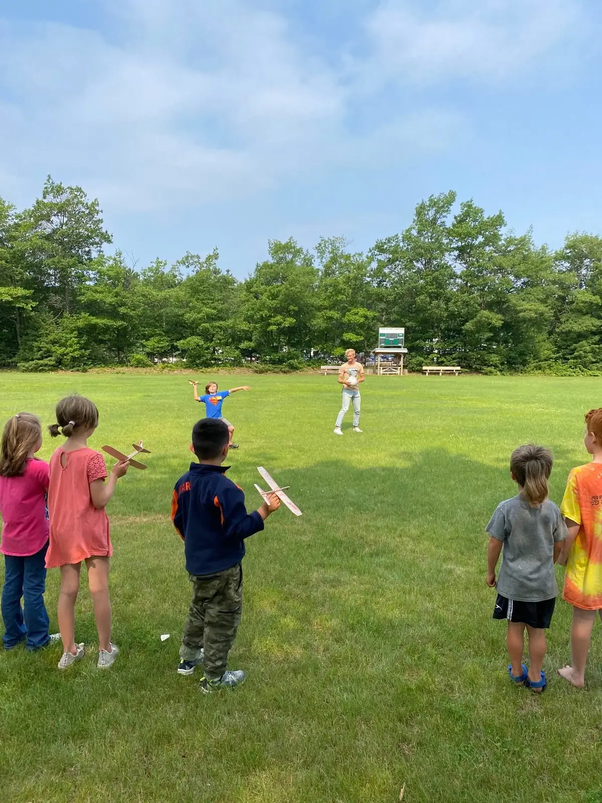 Children launch toy glider planes across a grassy field while an adult stands nearby.