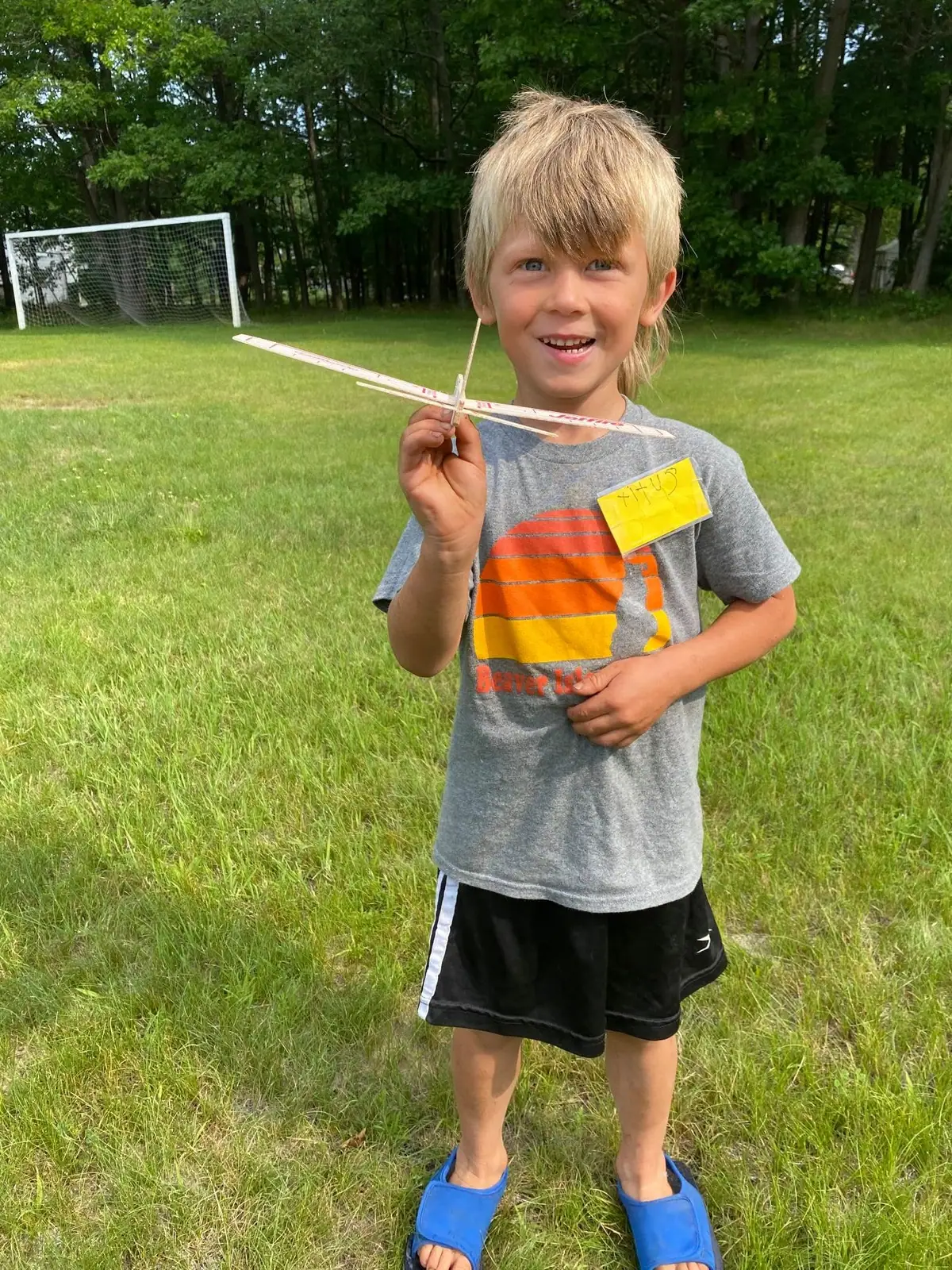 A child holds a small wooden model airplane in a grassy field beside a soccer goal.