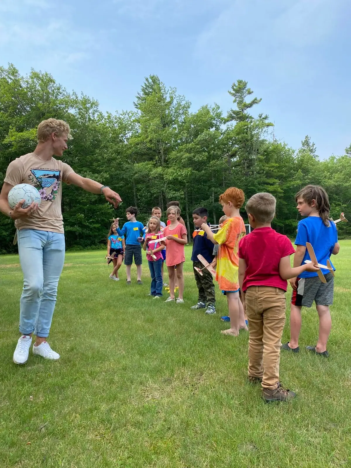 An adult holds a soccer ball and gestures toward children gathered with small wooden gliders on a grassy field.