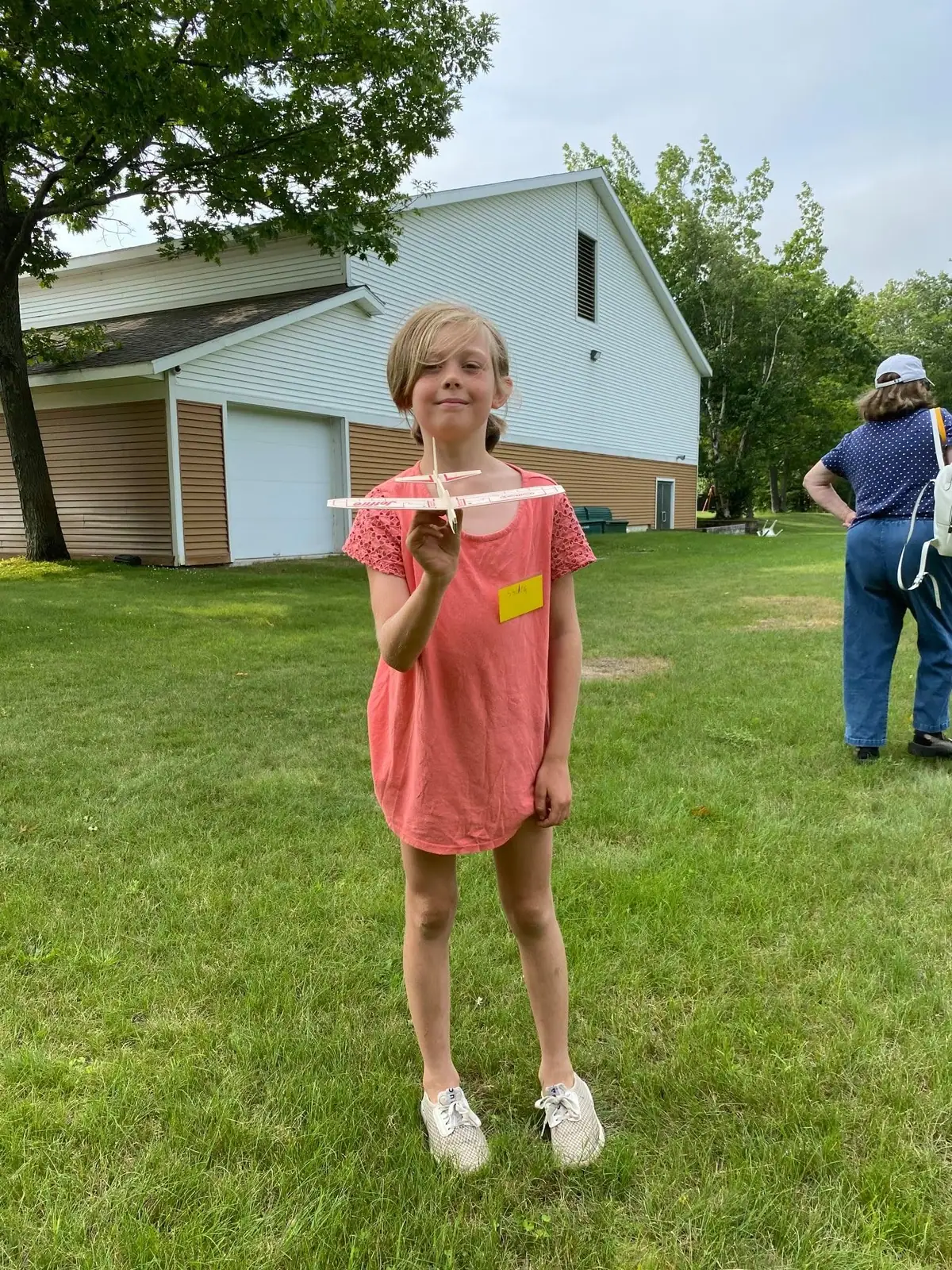 A child in a coral dress holds a model airplane on a grassy lawn beside a large wood-sided building.
