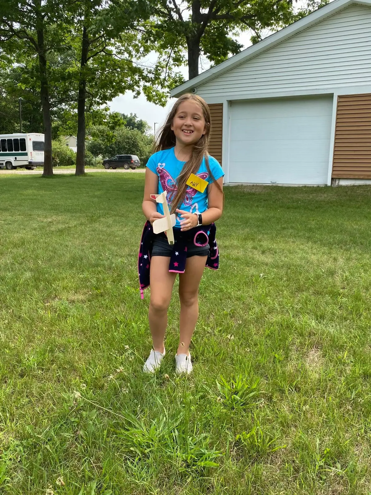 A smiling child holds a small wooden airplane model in a grassy yard beside a garage.