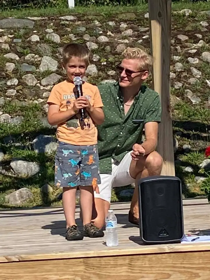 A child holds a microphone on a wooden platform beside a seated adult and a portable speaker.