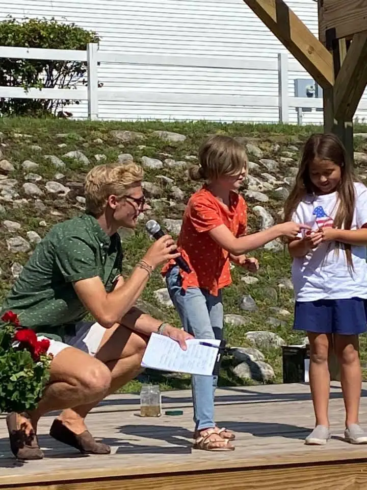 An adult holding a microphone and clipboard crouches beside two girls on an outdoor wooden platform.