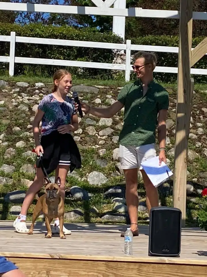 Two people speak into a microphone on a wooden stage beside a leashed dog.