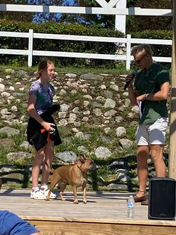 A girl and a man stand with a leashed dog on a wooden stage beside a microphone and speaker.