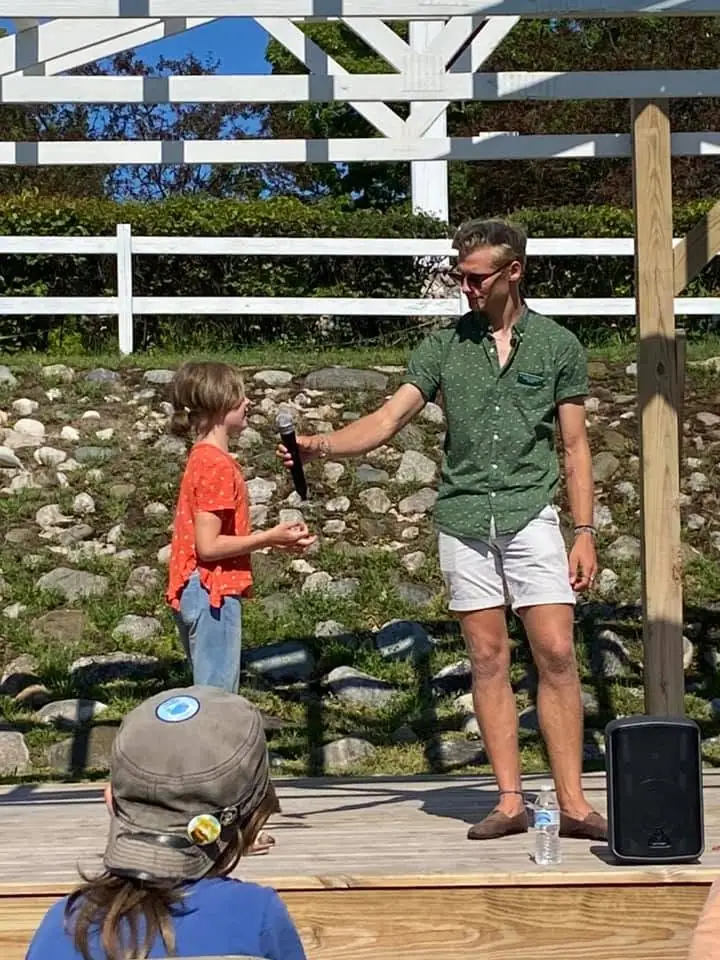 An adult holds a microphone toward a child on an outdoor wooden stage.