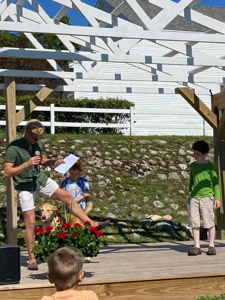 A man speaks into a microphone beside two children, a dog, and red flowers on a wooden stage.