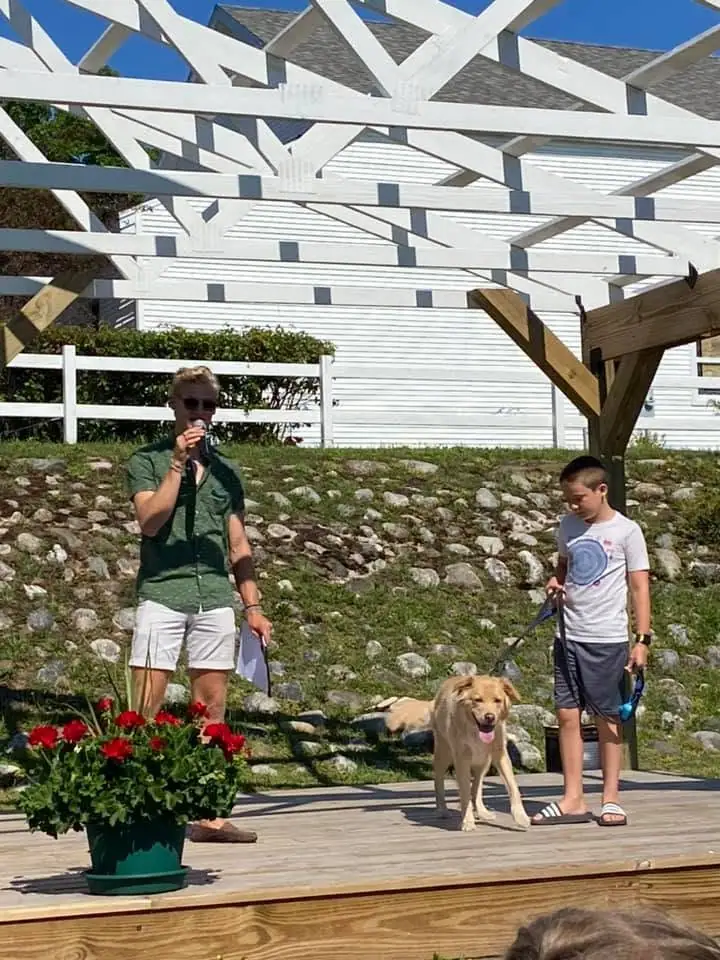 A woman speaks into a microphone beside a boy and dog on a wooden deck.
