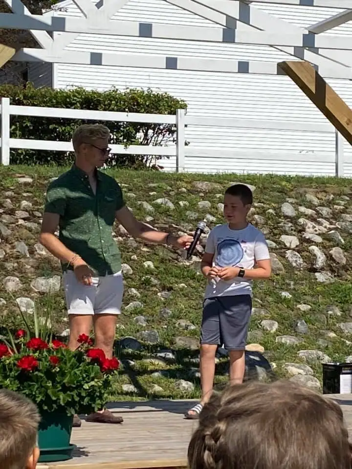 An adult holds a microphone toward a boy while several children watch from the foreground.