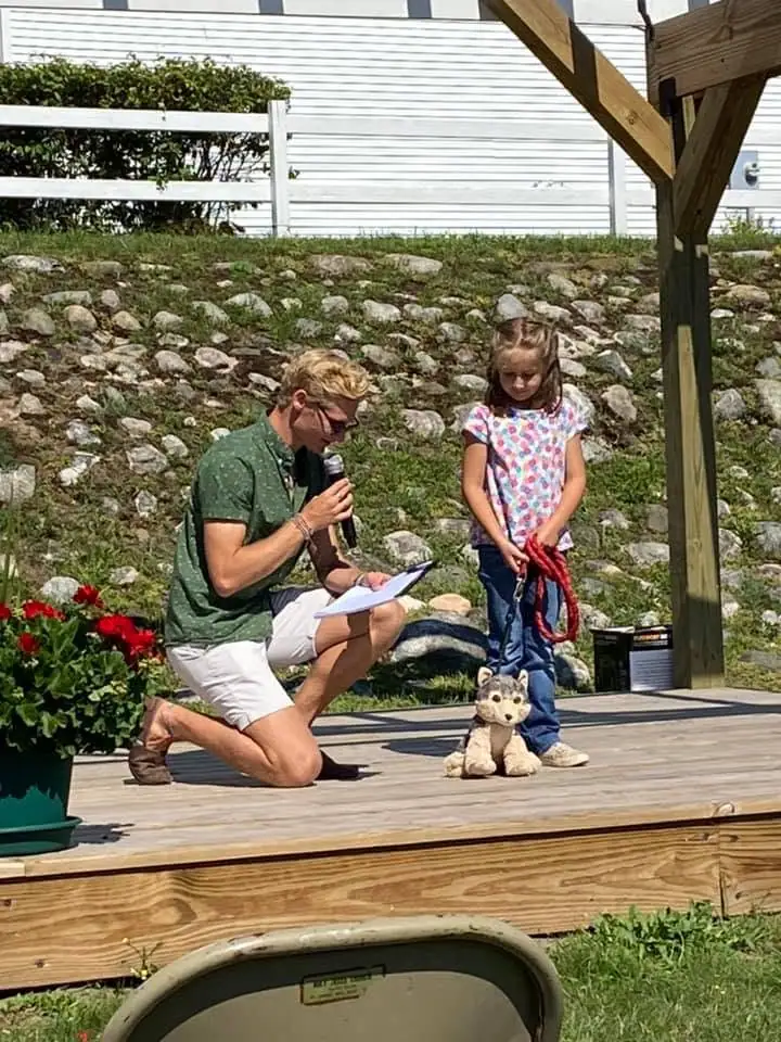 A man kneels with a microphone beside a child holding a red leash and a stuffed dog on a wooden stage.